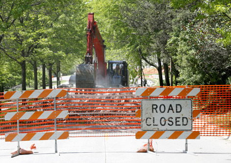 excavator digs up road surface behind "road closed" sign