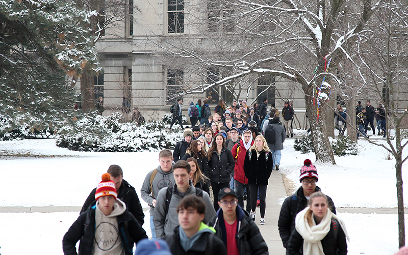 Students in caps and jackets move across a snowy campus lawn