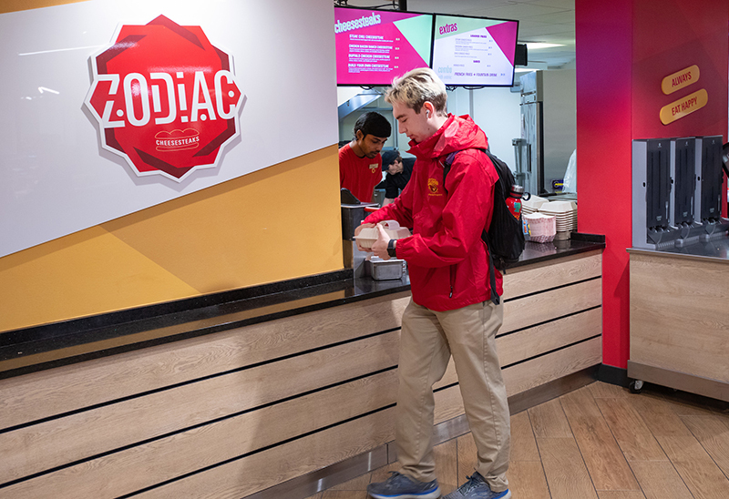 Male student in red jacket carries sandwich from food counter