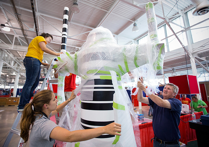 Three people remove plastic wrap from large metal sculpture