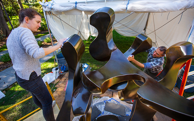 Two people apply clearcoat to large bronze sculpture