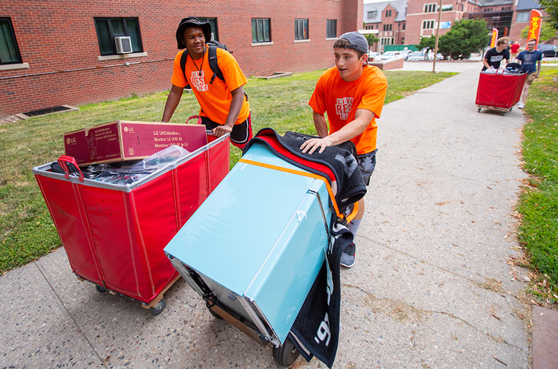 Two male students help move dorm room furnishings along the side