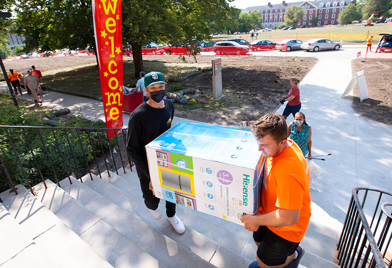 Two male students carry a boxed mini fridge up residence hall st