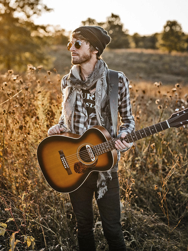 Joshua Sinclair holds his guitar in a field