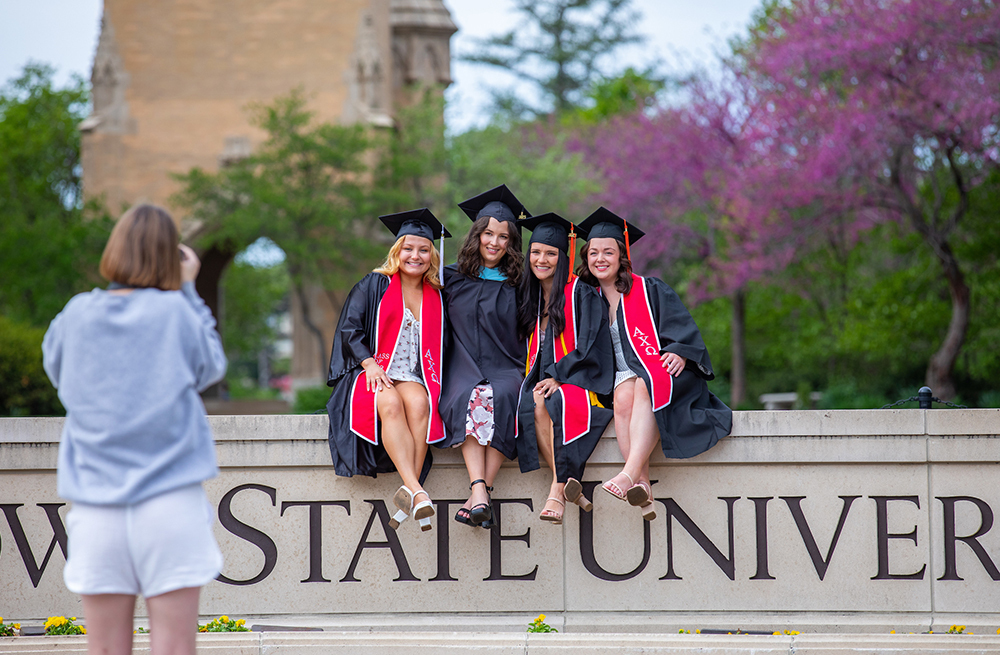 Four female students in graduation garb sit on the ISU wall