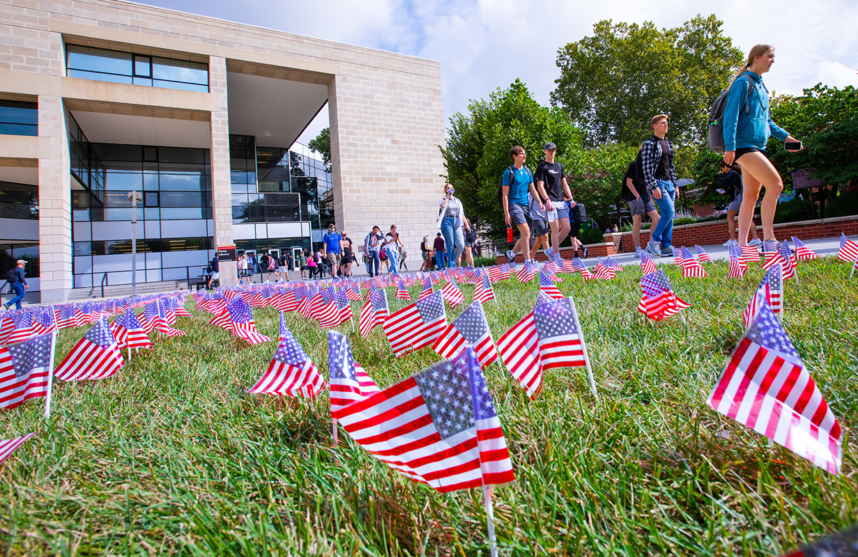 Students walk by rows of small American flags stuck in a grass l