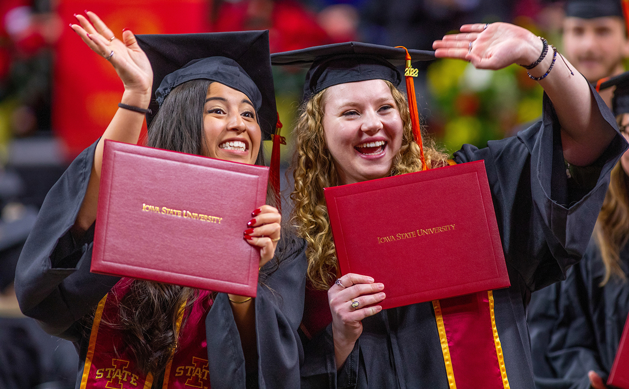 Female students in academic robes smile and hold their red diplo